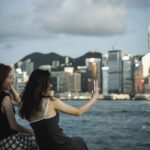 Niezbędne aplikacje na telefon w Hongkongu: transport, płatności, mapy i zamawianie jedzenia Dwie kobiety robią selfie przy porcie i panoramie Hongkongu